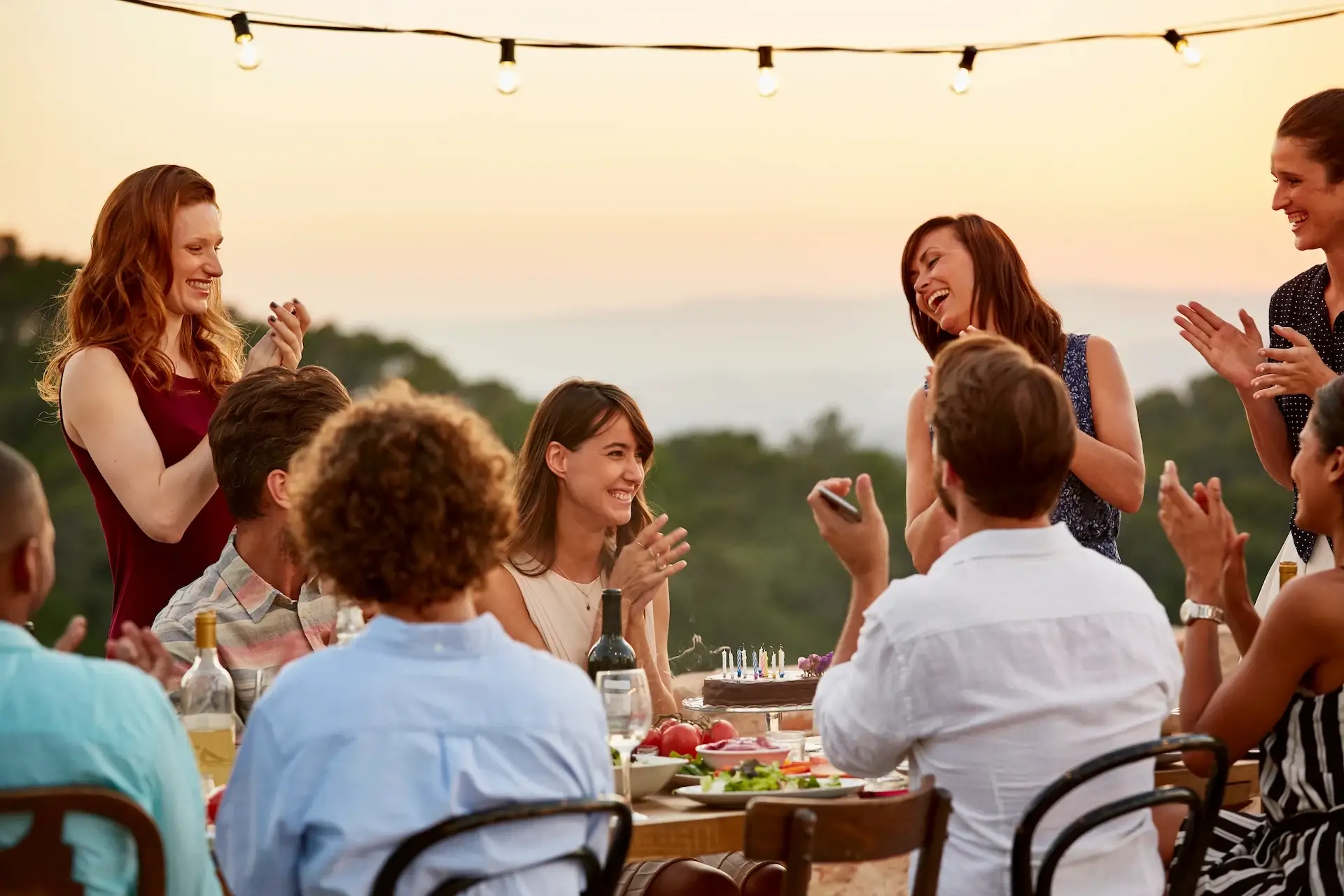 Group of friends joyfully celebrating a birthday at an outdoor table, with plenty of food and drinks.
