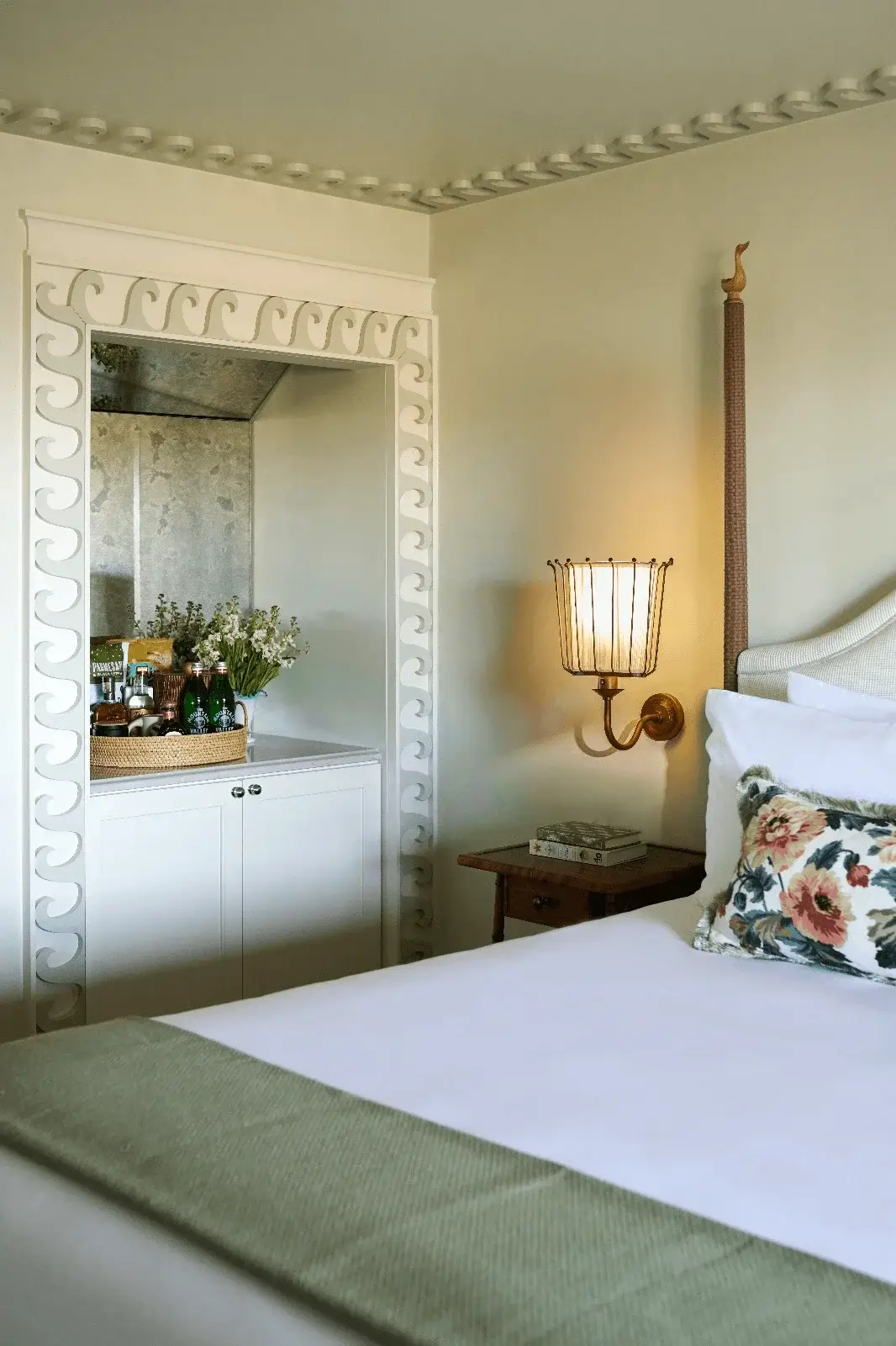 King-sized bed at The Sanderling Resort next to a nightstand with a lit lamp, and a dresser with fresh flowers and drinks.