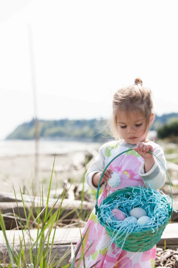 A little girl on the beach with a green basket, collecting painted eggs, surrounded by a beautiful landscape.