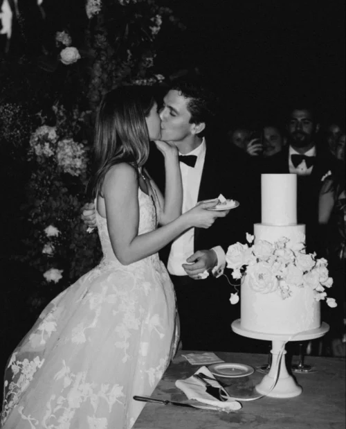 Black and white photo of a young newlywed couple kissing next to their wedding cake.