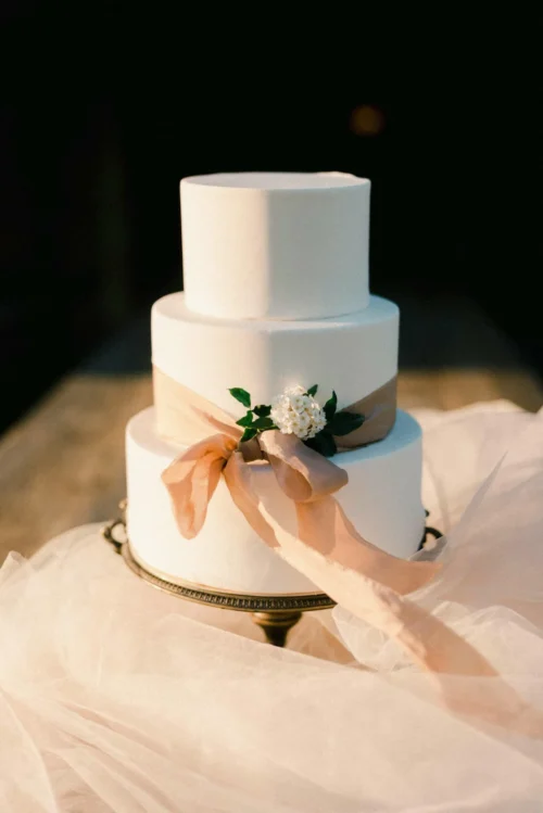 Wedding cake on a copper base with a bow and white flowers, placed on a wooden table.